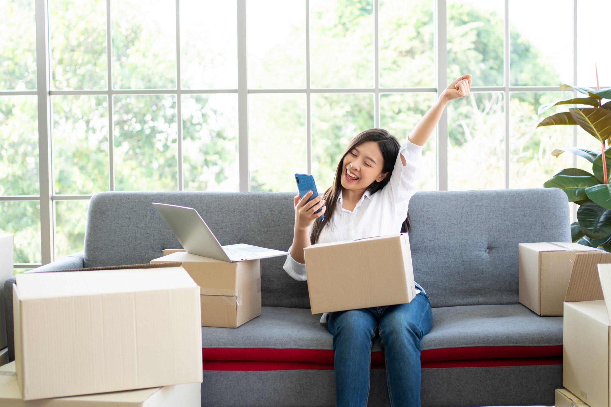 Happy Young Asian Woman Entrepreneur, Smile for Sales Success after Checking Order from Online Shopping Store in a Smartphone at Home Office, Concept of Merchant Business Online and ECommerce